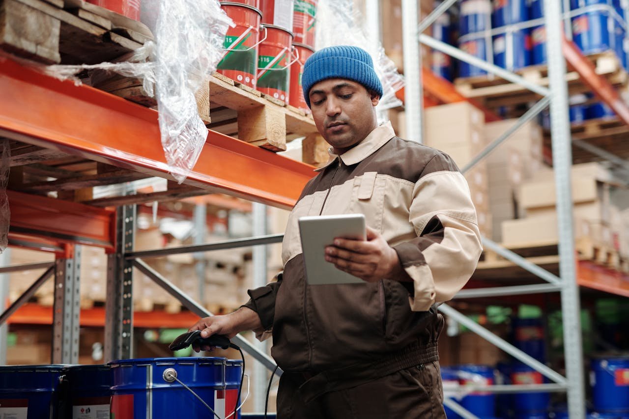 Contact Man in blue beanie uses tablet in a warehouse for inventory management.